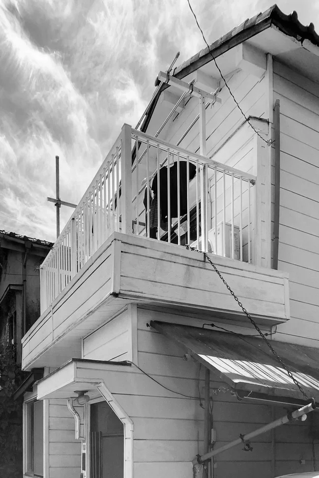 白黒で撮影された住宅のバルコニーと外観。白い手すりのあるバルコニーには洗濯物のようなものが見える。曇り空の下、建物の構造や生活感が伝わる日常の一コマ。