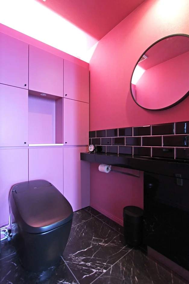 ピンクと黒を基調としたモダンなトイレルームのインテリアです。黒いタンクレストイレ、大理石調の床、収納棚、丸い鏡が配置されており、個性的でスタイリッシュな空間を演出しています。