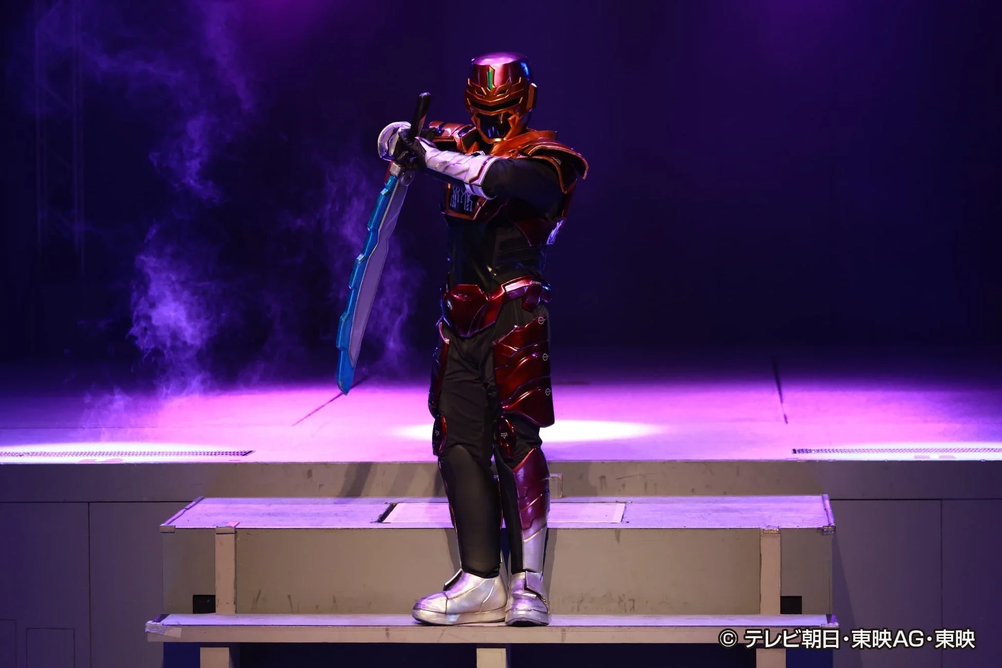 赤いアーマーを身につけたヒーローのようなキャラクターが、剣を構えて紫色の照明と煙が演出されたステージに立っています