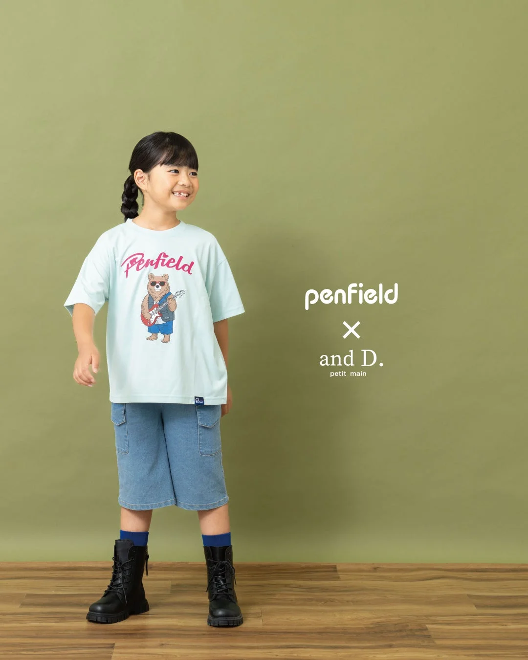 笑顔の子供がPenfieldのクマのイラストTシャツ、デニムのハーフパンツ、黒いブーツを着用