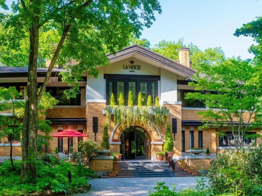 緑豊かな自然に囲まれた「LA NEIGE HOTEL」の美しい外観。レンガ調の建物と手入れされた庭園が調和し、晴れた日の光の中で落ち着いた高級感を醸し出しています。