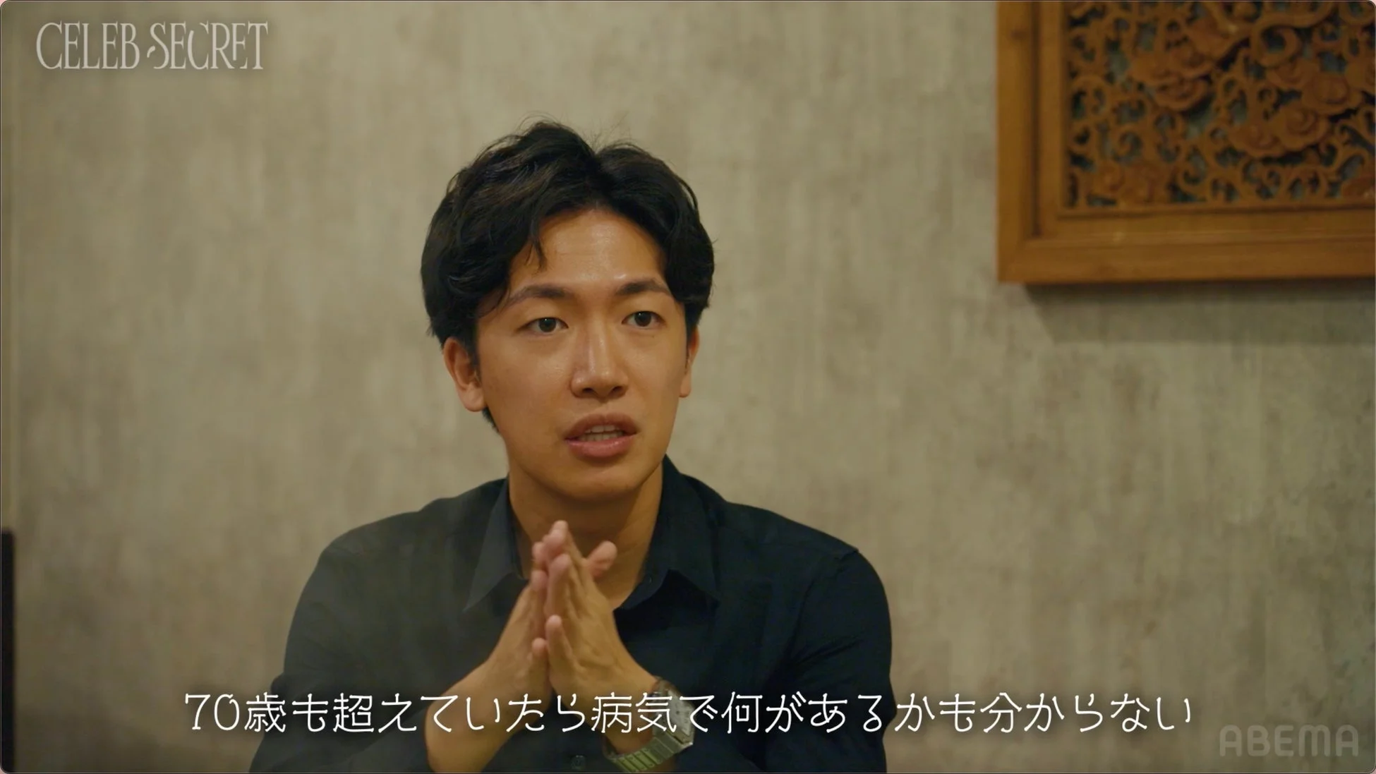 室内で若い男性が真剣な表情で話しており、両手を組んでいます。