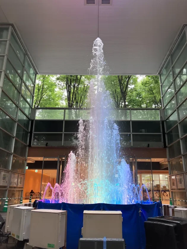 商業施設のようなモダンな建物内で、虹色にライトアップされた噴水が力強く水を噴き上げている