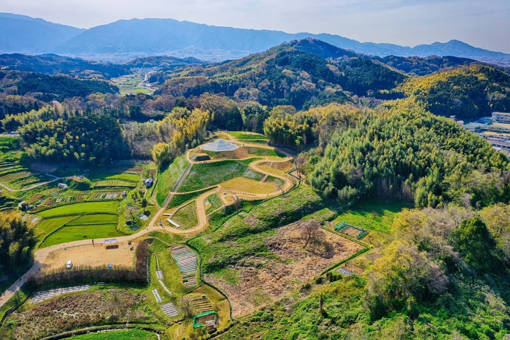 空撮された、緑豊かな山々と段々畑が広がる自然豊かな風景。丘の上には特徴的な円錐形の建物が見え、整備された小道が続く。