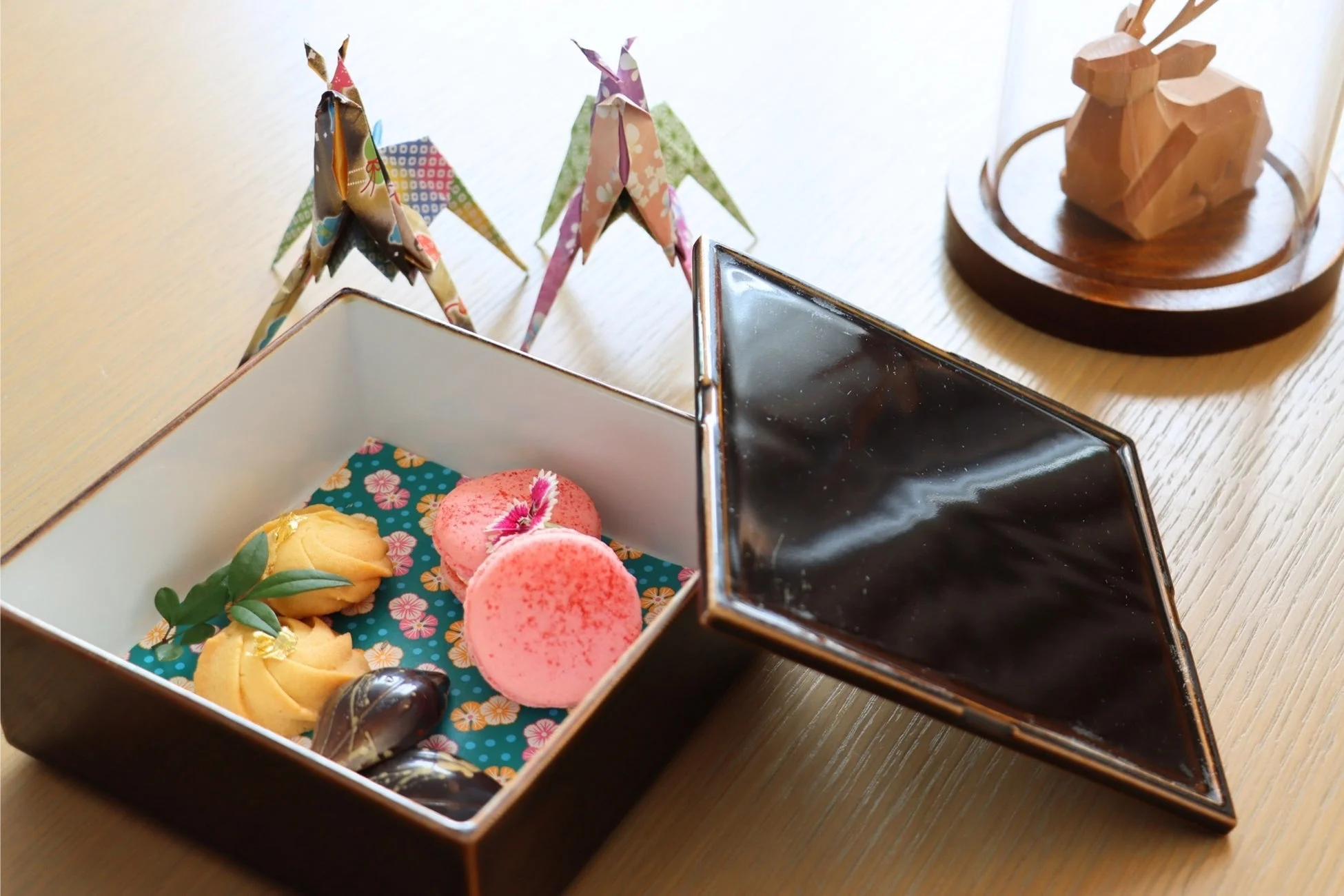 華やかな和柄の布が敷かれた箱に、マカロン、クッキー、チョコレートなどの様々なスイーツが美しく盛り付けられている。背景には折り紙の飾りや木製の鹿の置物があり、上品で可愛らしい雰囲気を演出している。