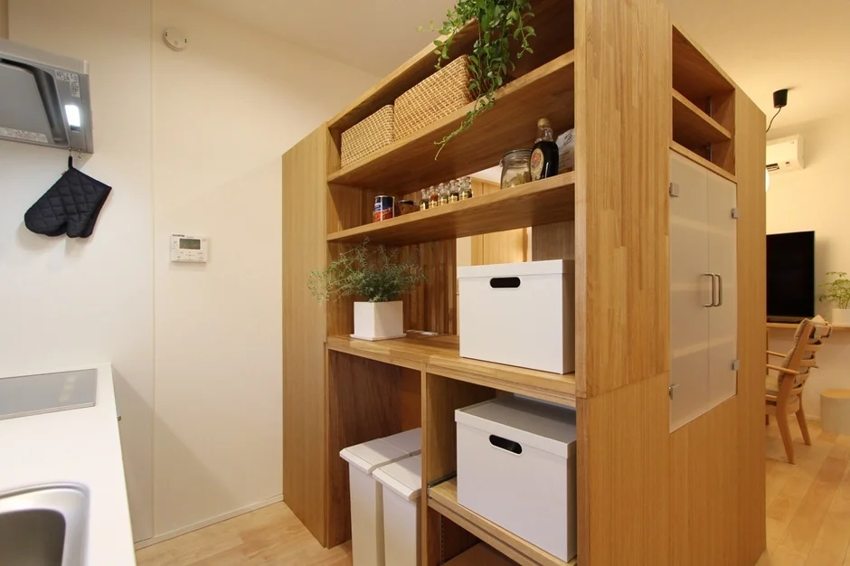 木製の収納棚で仕切られた、キッチンとリビングが一体となった機能的な空間。観葉植物や収納ボックスで整理整頓されており、ナチュラルで整った印象のインテリアです。