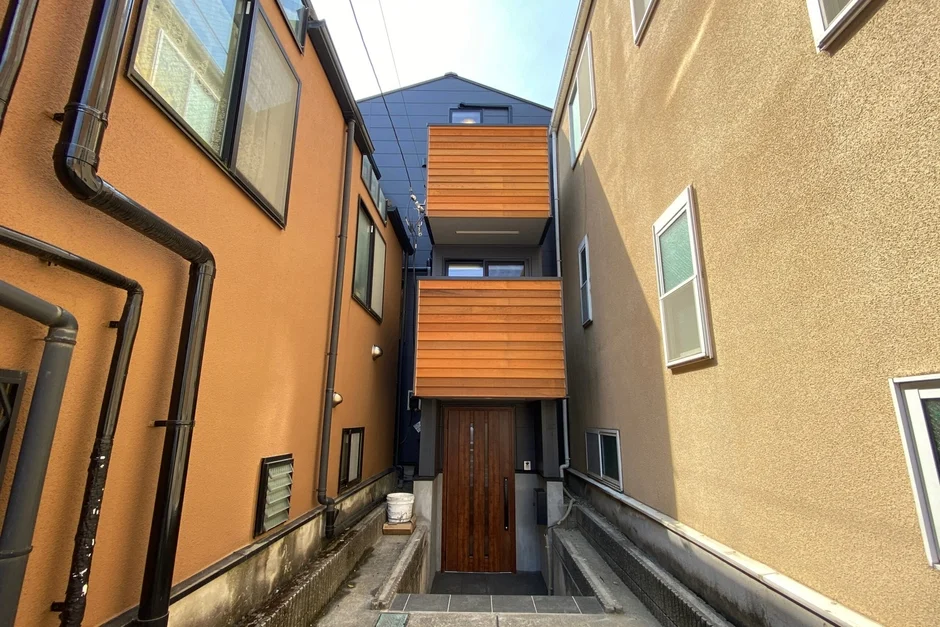 狭い路地の奥に位置するモダンなデザインの住宅。木製バルコニーが特徴的で、両側の建物に挟まれた独特の空間が広がっています。