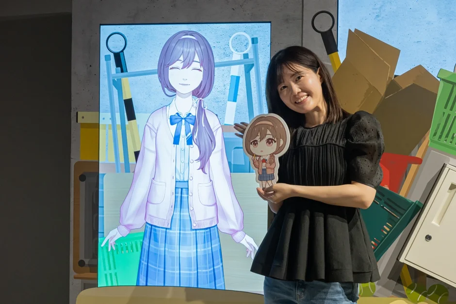 ディスプレイに映るアニメキャラクターとミニキャラクターグッズを持つ長妻樹里さん