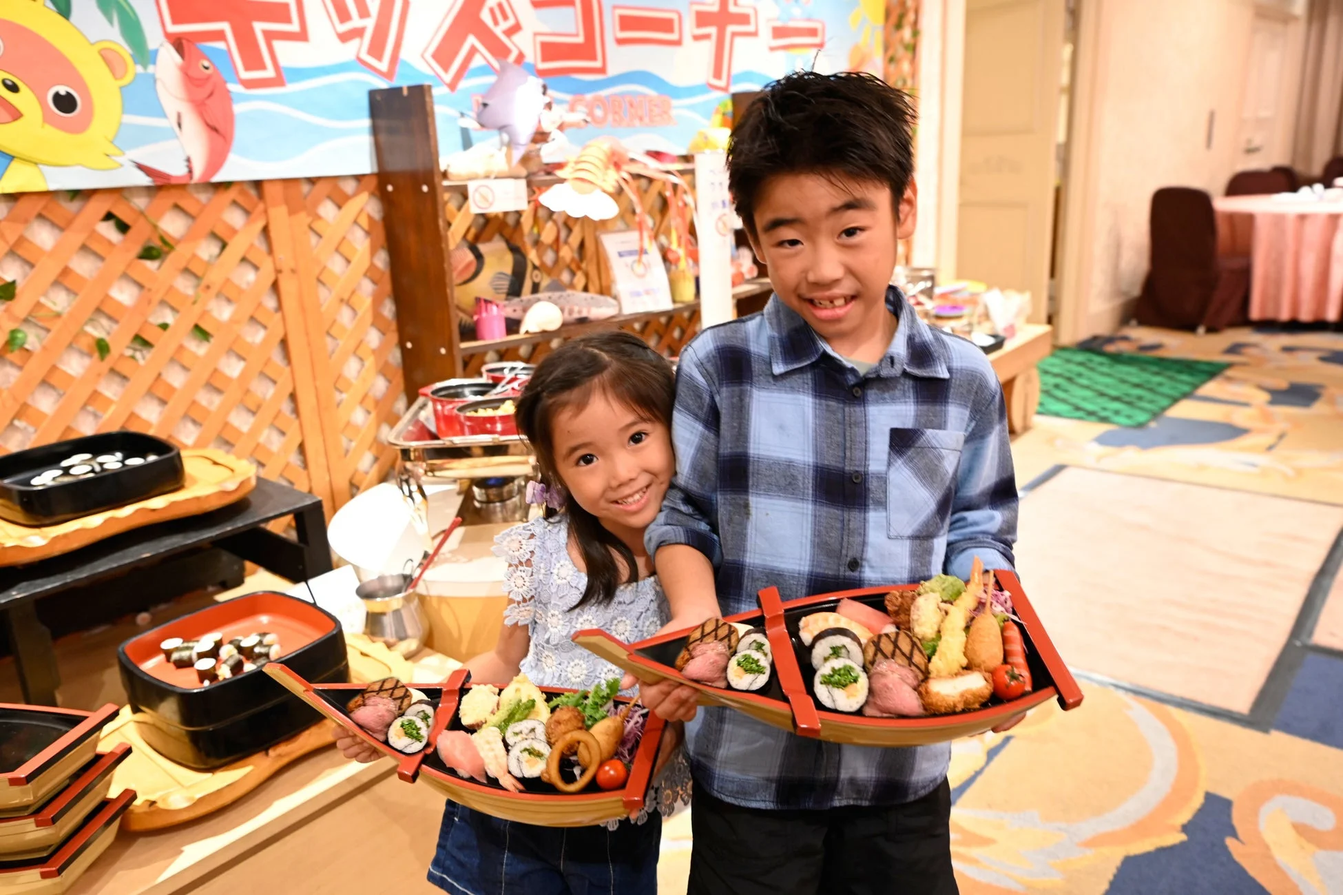 キッズコーナーで、男の子と女の子が笑顔で船盛りの寿司や天ぷらなどの和食を持っている様子