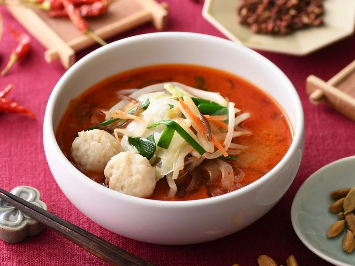 赤い食卓に置かれた白い器に入った辛そうな中華スープ。肉団子、キャベツ、もやし、春雨などの具材が豊富で、背景には唐辛子や香辛料が見える。食欲をそそる一品。
