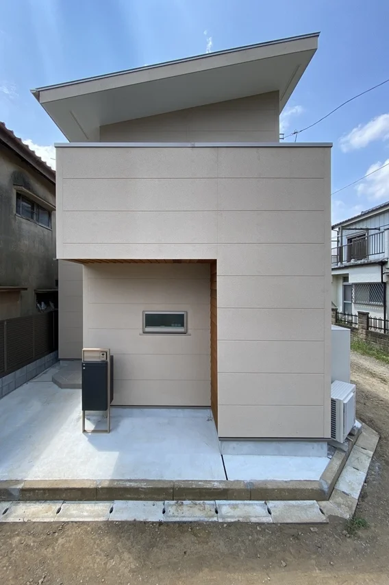 傾斜した屋根が特徴的な、ベージュ色の現代的な一軒家の外観です。シンプルなデザインで、玄関付近にはポストと小さな窓が見え、青空の下に建っています。