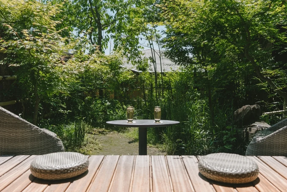 豊かな緑に囲まれた和風の庭園で、ウッドデッキに置かれたテーブルと椅子に飲み物が並んでいます。