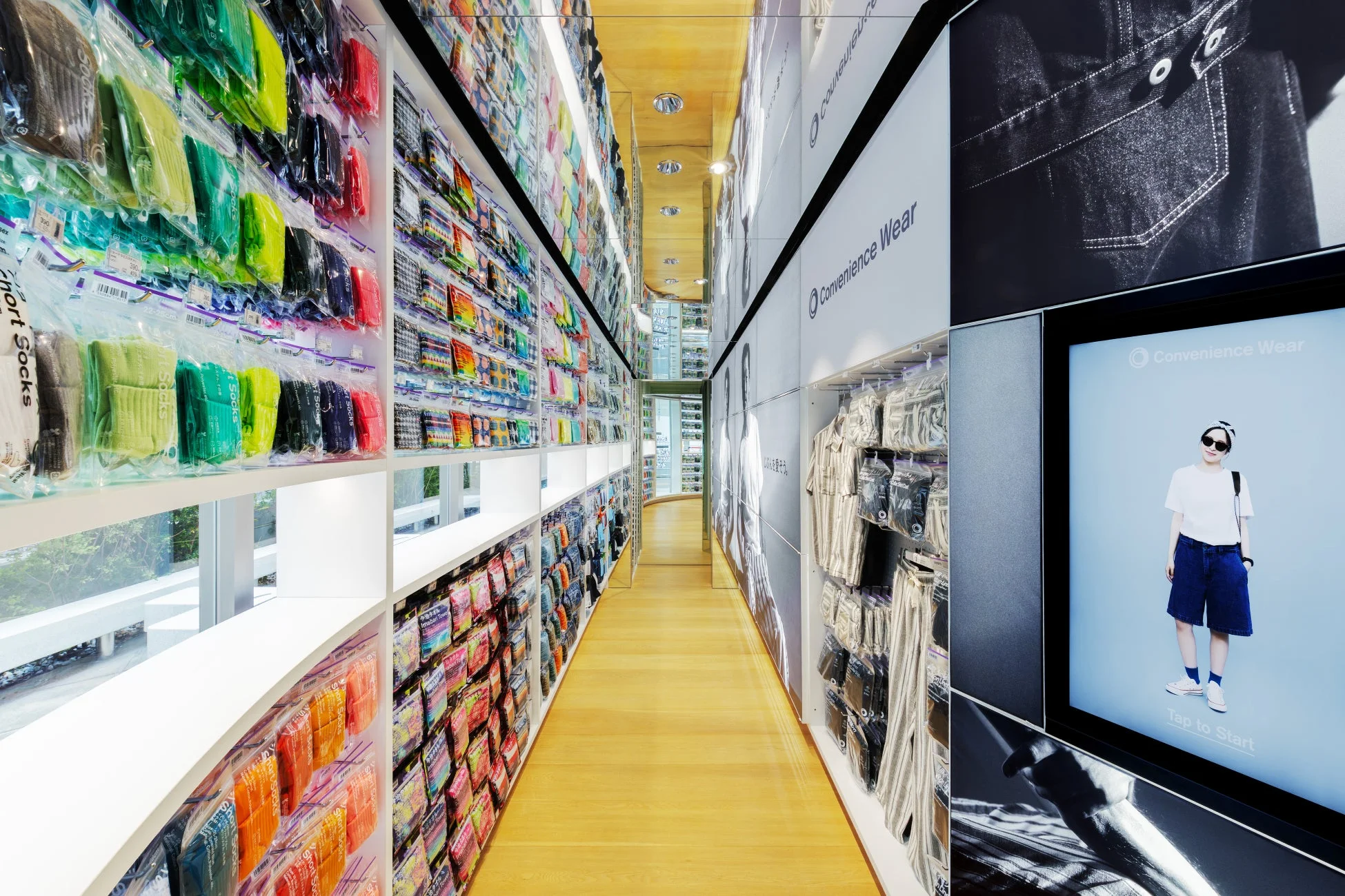 モダンなアパレル店舗の通路で、カラフルな靴下や衣料品が棚に陳列されています。右側の壁には「Convenience Wear」の商品が並び、デジタルサイネージにはモデルの画像と「Tap to Start」の文字が表示されています。