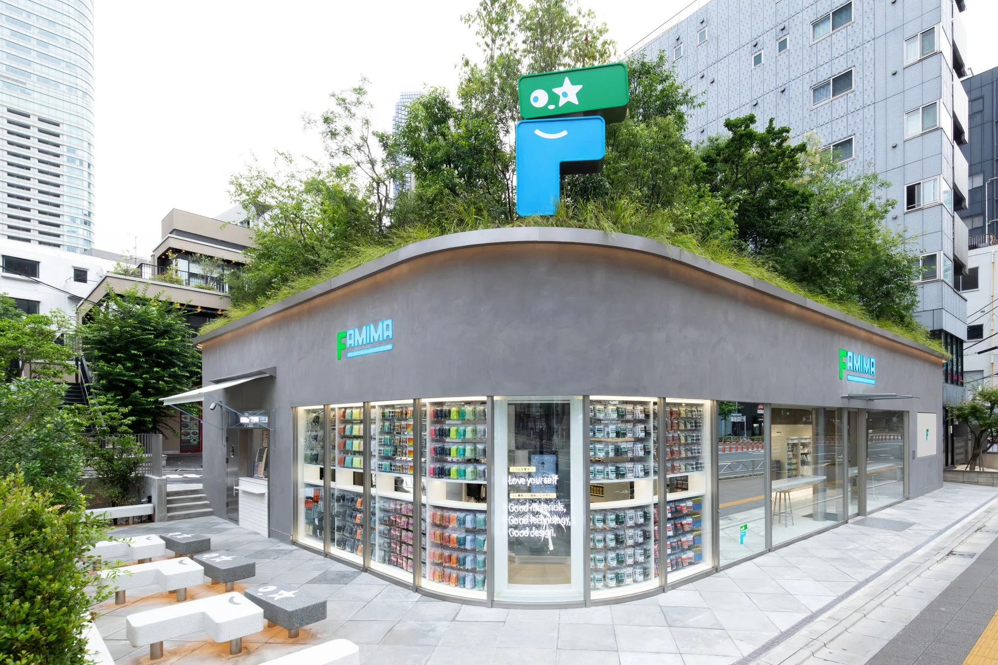 都会的な環境に溶け込む、緑化された屋根が特徴のモダンな「FAMIMA」コンビニエンスストア。ユニークなロゴとガラス張りの外観を持ち、店内には様々な商品が並び、屋外にはデザイン性の高いベンチが設置されている。