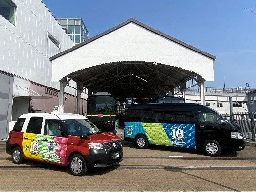 記念デザインが施された赤いタクシーと黒いバンが、車庫のような場所で停車している様子。奥には緑色の電車も見え、晴れた日の屋外で撮影された写真です。