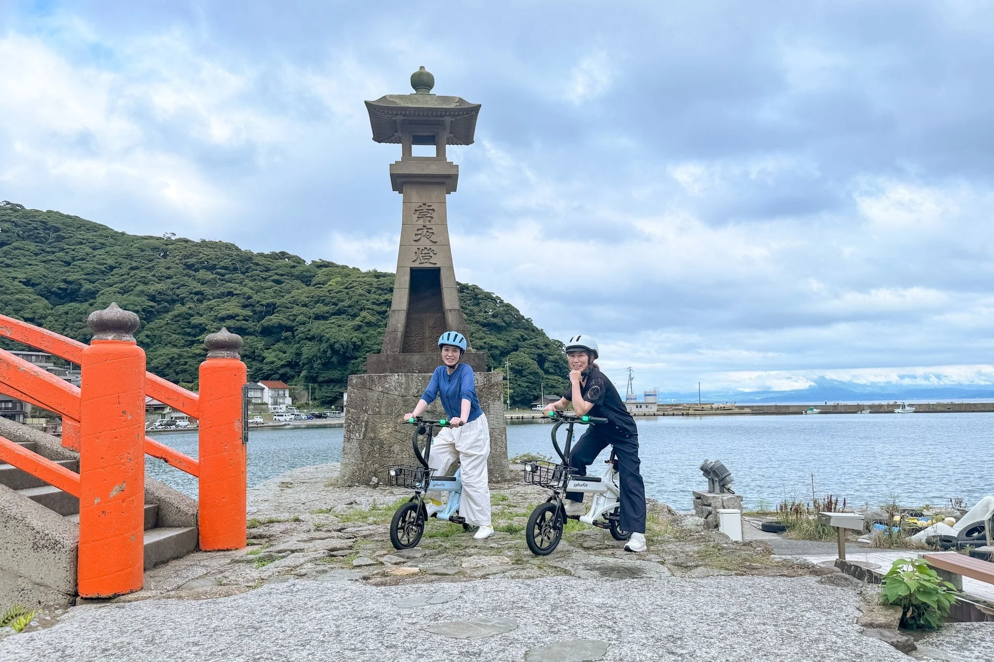女性二人、電動自転車、石灯籠、港、海、観光、旅行、サイクリング、橋、山、曇り空