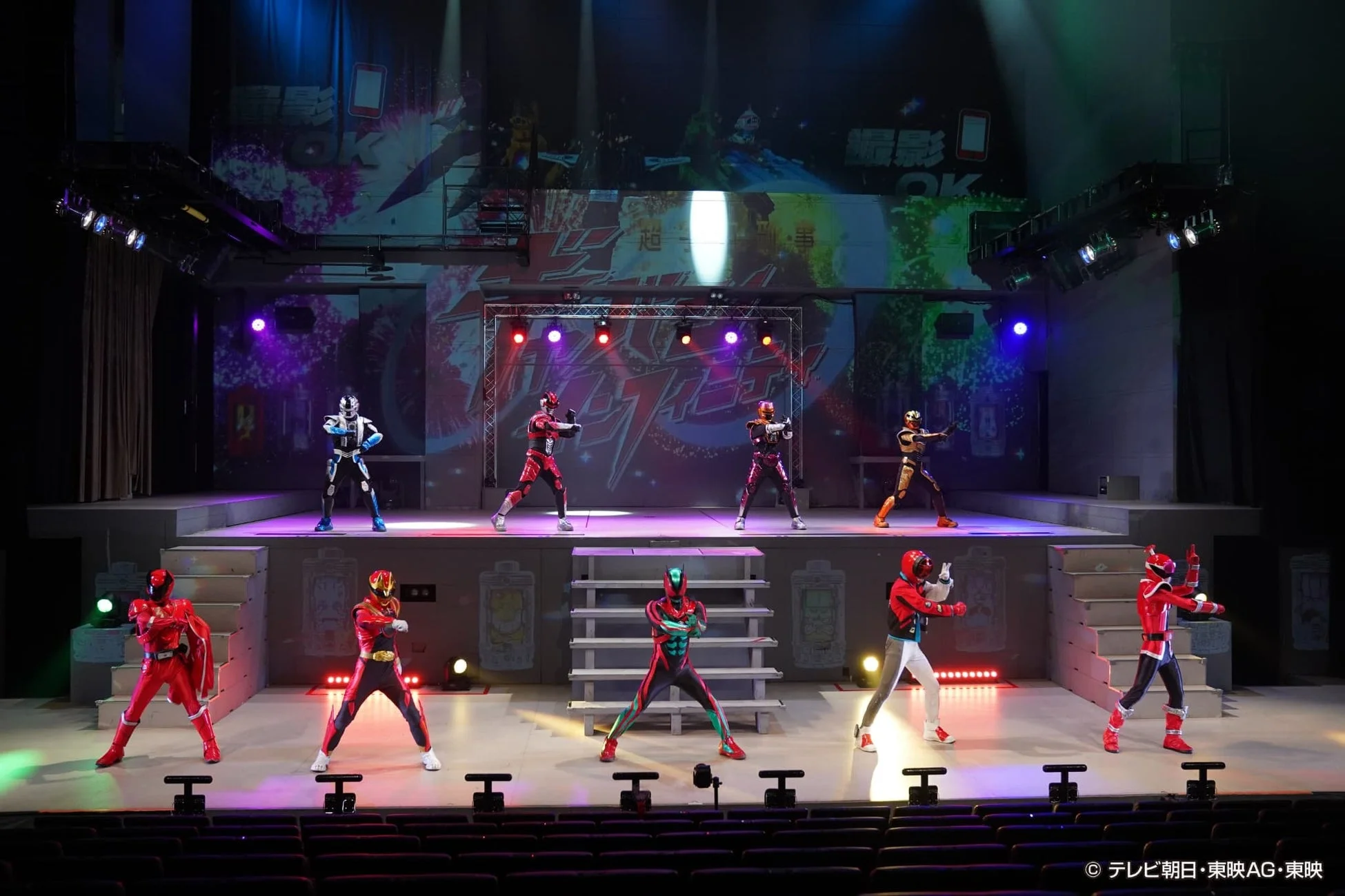 特撮ヒーローのライブステージパフォーマンス。カラフルなコスチュームをまとった複数のヒーローたちが、照明が輝く多層ステージでポーズを決めている。