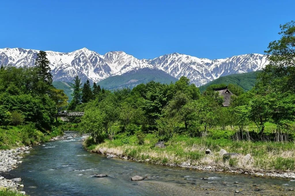 雪を頂いた雄大な山々を背景に、清らかな川が流れ、豊かな緑に囲まれた日本の美しい風景。茅葺き屋根の家屋も見える。