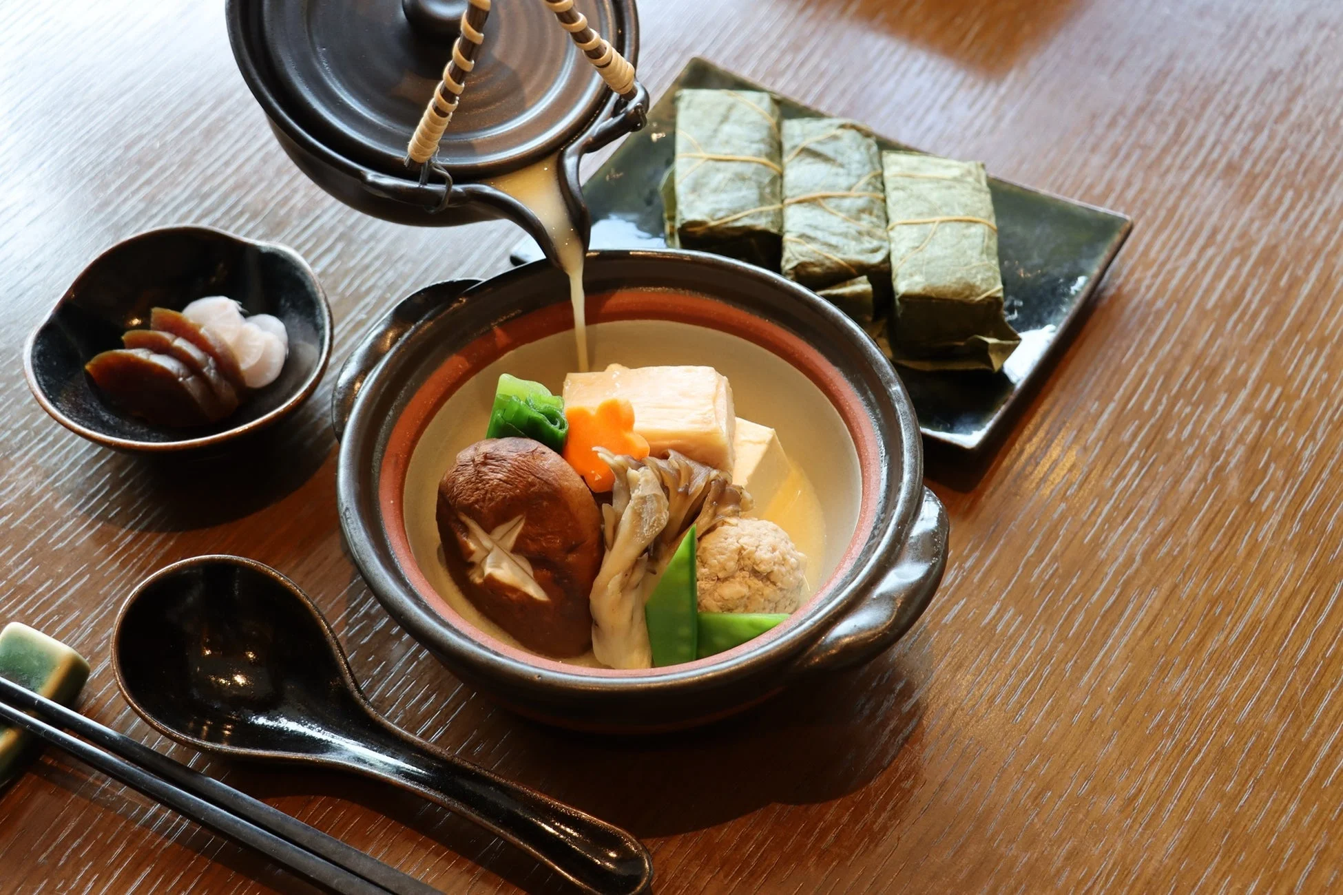 木のテーブルに置かれた和食の膳。土鍋風の器に豆腐やきのこ、野菜などの具材が入った煮物が盛られ、上から出汁が注がれている。笹に包まれた料理や漬物も添えられ、彩り豊かで美味しそうな食卓の様子。