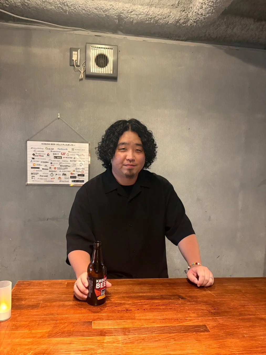 黒いシャツを着たパーマヘアの男性が、木製のカウンター越しにKOBUSHI BEERのボトルを持ち、微笑んでいる。背景には「KOBUSHI BEER コミュニティスポンサー」と書かれたボードが見えるバーのような空間。
