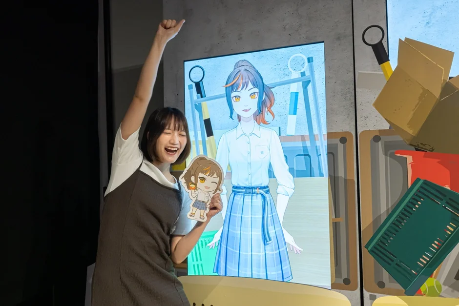 笑顔でポーズをとる菱川花菜さんとバーチャルキャラクターのディスプレイ
