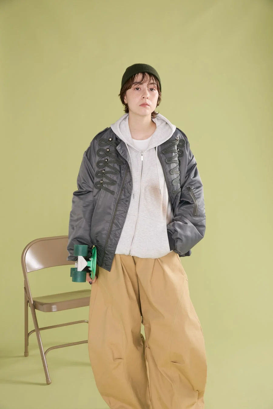 緑のビーニーとグレーのボンバージャケットを着用し、ベージュのワイドパンツを合わせた女性の全身ポートレート。