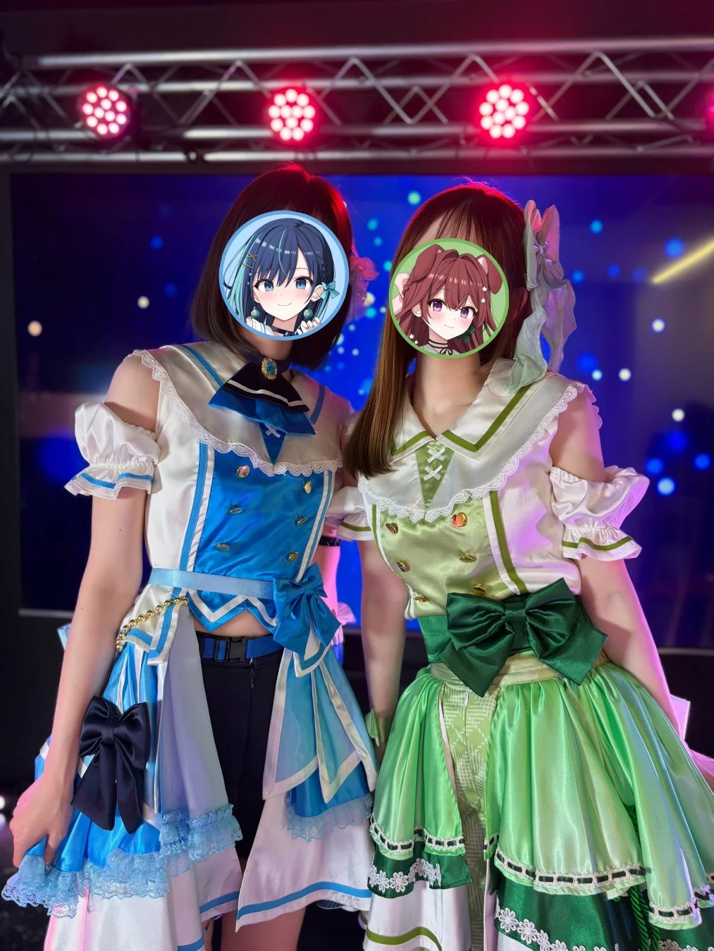 ステージ上でポーズをとる2人の女性の姿。それぞれ青と緑を基調とした華やかなアイドル衣装を着用しており、顔はアニメキャラクターのイラストで隠されている。背景にはステージ照明と大きなスクリーンが見える。