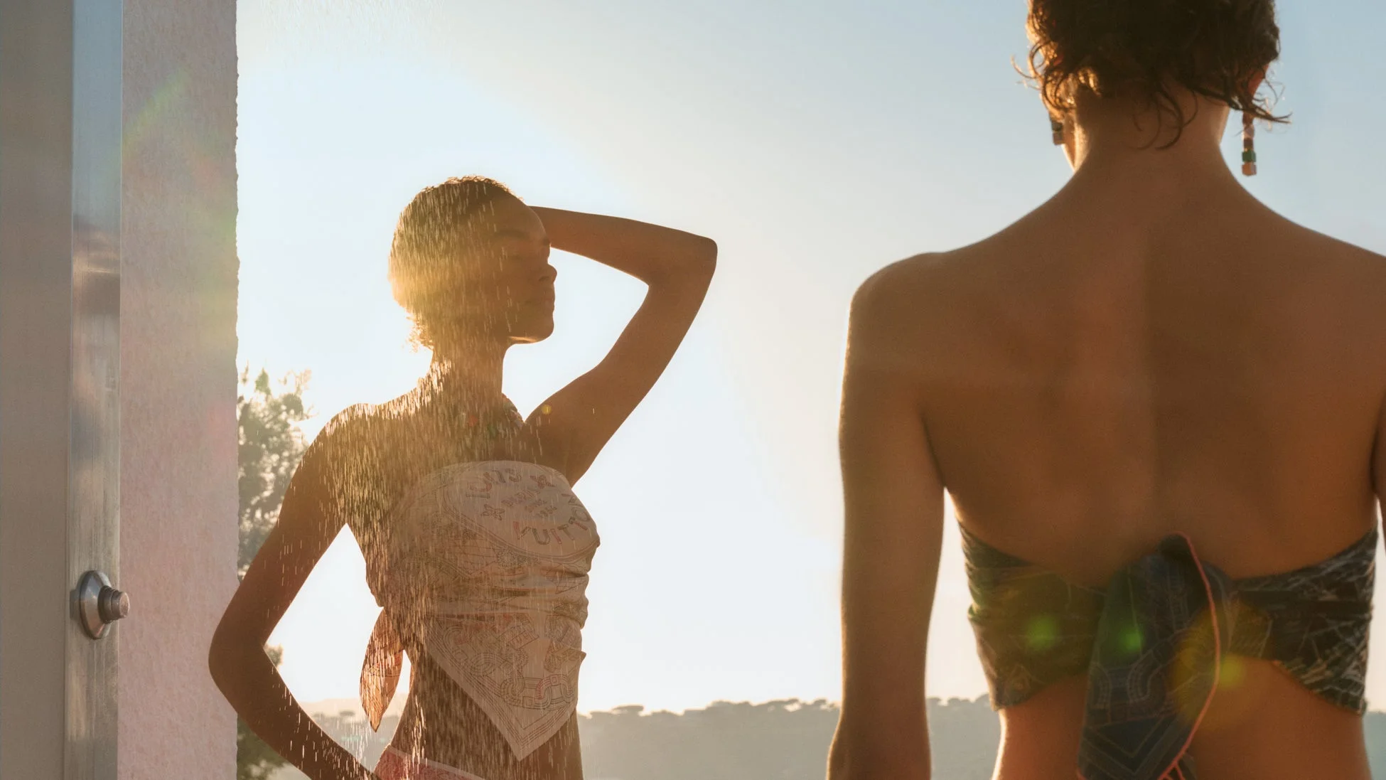 夕日の逆光の中でシャワーを浴びる女性と、後ろ姿の女性が写る。ルイ・ヴィトンのスカーフを着用し、リゾート地でのリラックスした夏のひとときを捉えた写真。