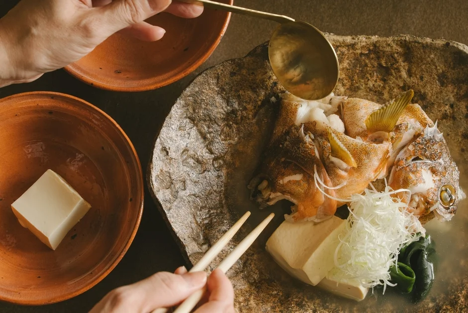 温かい土鍋で煮込まれた魚料理を中心に、豆腐や薬味が添えられた和食の食卓風景です。