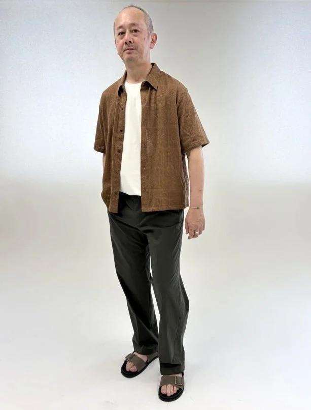 白いTシャツの上に茶色の半袖シャツを羽織り、ダークカラーのパンツとサンダルを着用した男性
