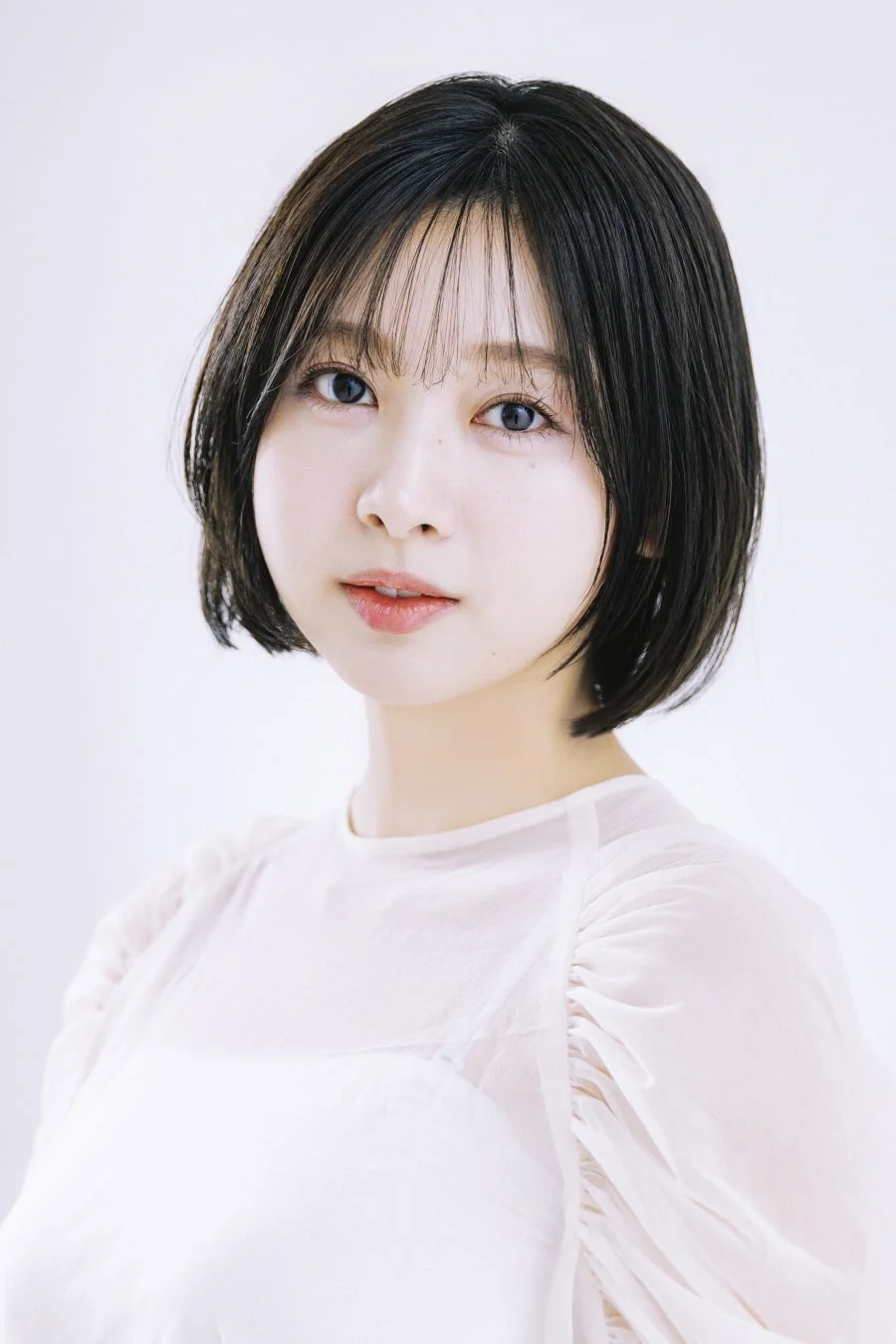 白い背景の前で、白いシースルーのトップスを着たショートヘアの若い女性のポートレート。彼女はカメラを見つめ、穏やかな表情をしています。