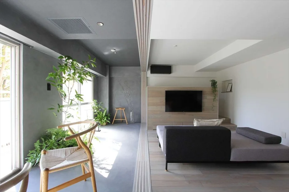 大きな窓から光が差し込むモダンなリビングルーム。観葉植物が豊かな空間と、ウッドパネルの壁にテレビが配された落ち着いたエリアが融合しています。