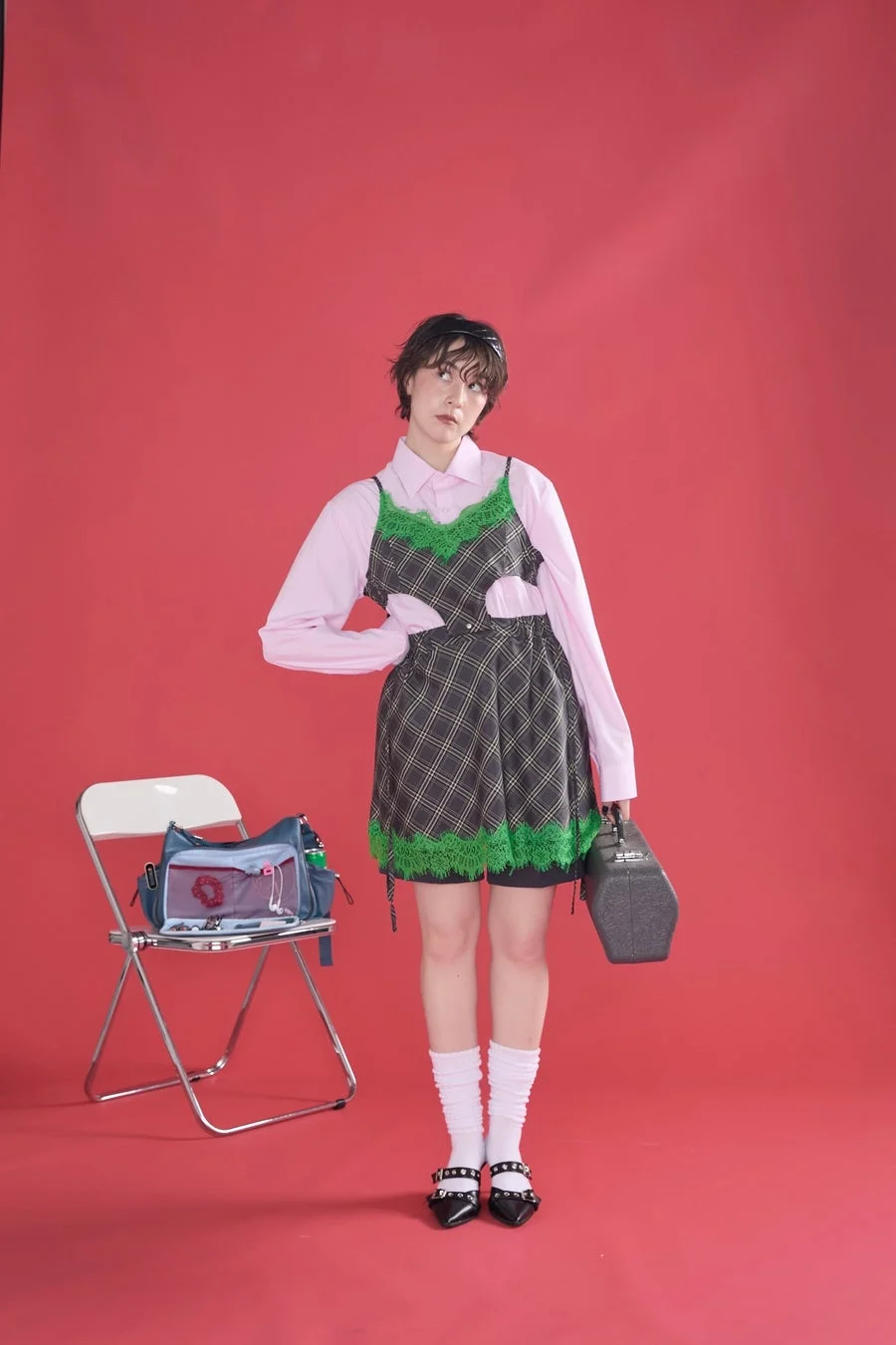赤い背景の前で、ピンクのシャツに緑色のレースがあしらわれたチェック柄のワンピースを重ね着した女性がポーズをとっています。