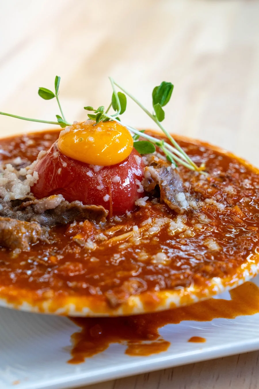 丸ごとトマトと鮮やかな卵黄が目を引く、赤い濃厚ソースの料理です。薄切り肉と白い粒状の食材がソースに絡み、緑の芽が彩りを添えています。器から溢れるソースが食欲をそそる一皿です。