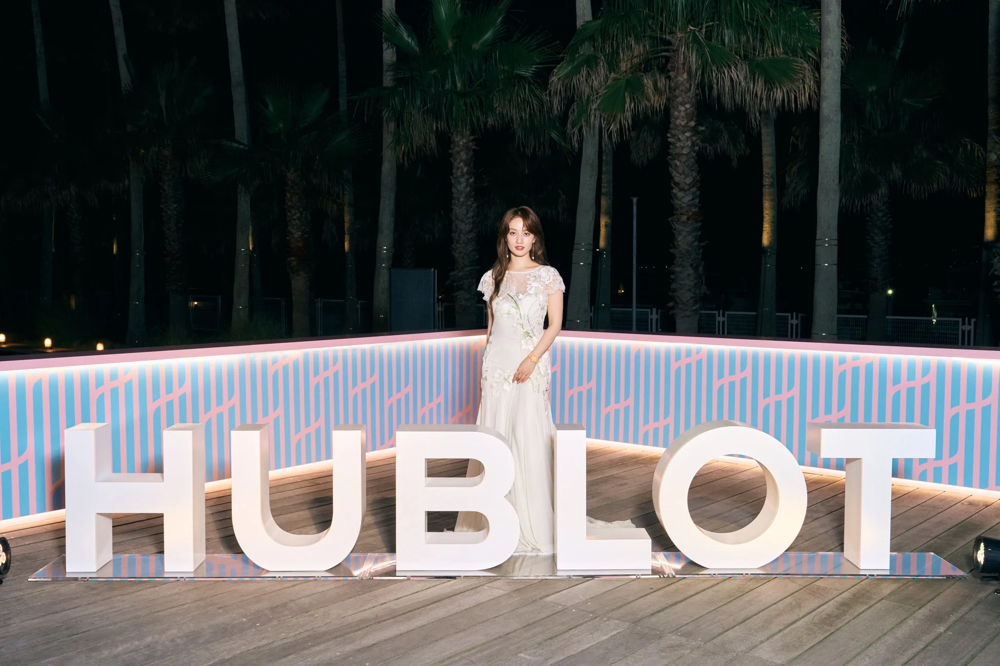 白い花柄のドレスをまとった女性が、カラフルなストライプの背景と「HUBLOT」の巨大なロゴの前に立っています