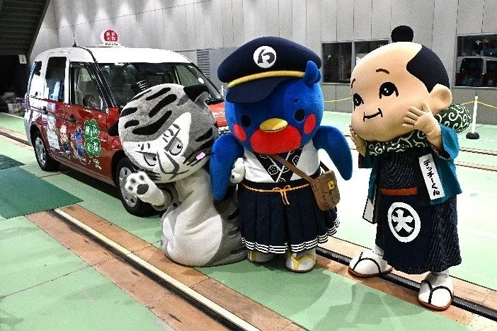 観光タクシーの横に、トラ、鉄道員風の鳥、和装の少年「デッチーくん」の3体のゆるキャラが並んでいるイベント風景です。