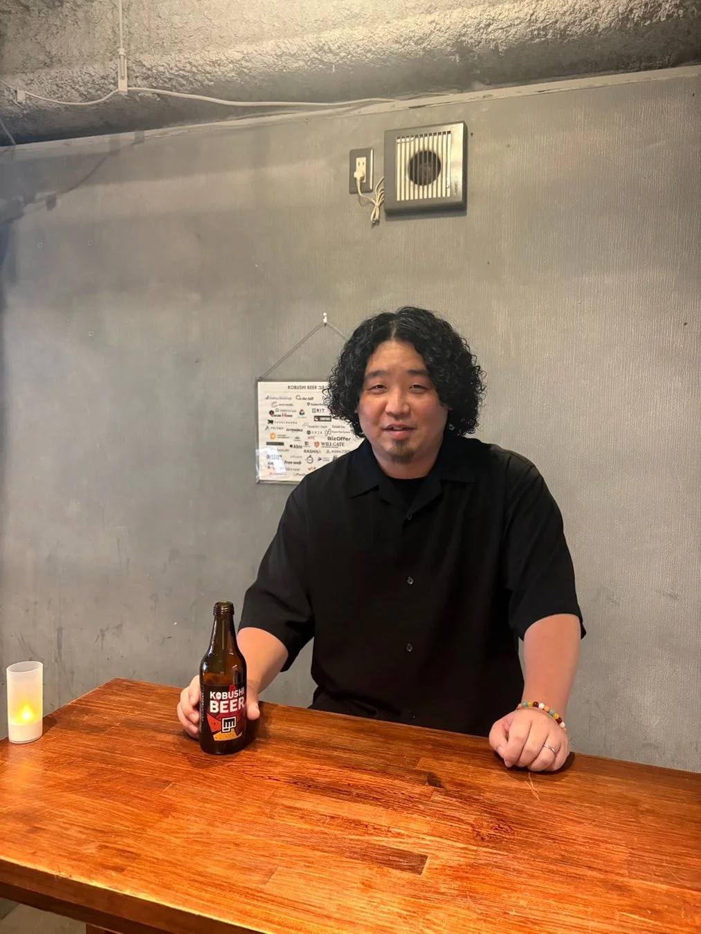 木製カウンターで黒いシャツの男性が笑顔でKOBUSHI BEERのボトルを持っている。背後には広告ポスターが見え、バーのような雰囲気の写真。