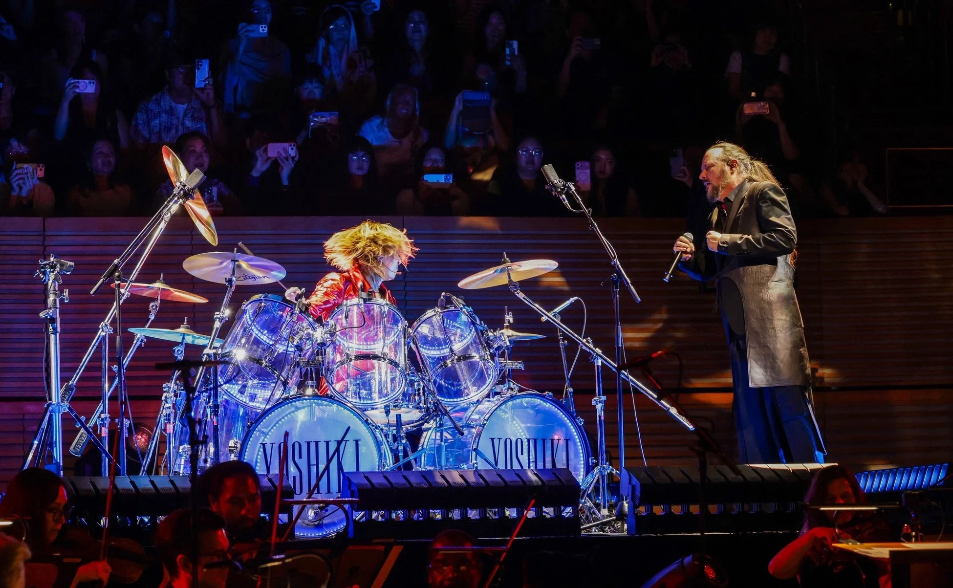 YOSHIKIが透明なドラムセットを演奏し、別の男性ヴォーカリストが歌唱しているライブパフォーマンスの瞬間を捉えた画像です。観客が携帯電話で撮影しており、オーケストラのメンバーも手前下部に写っています。ステージは青い照明で照らされています。