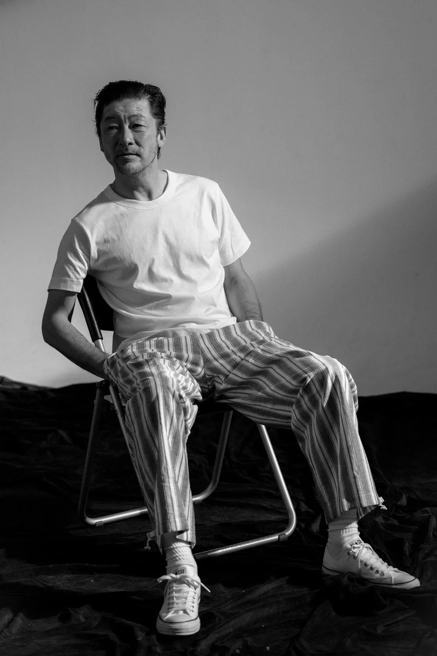 白黒写真で、椅子に座った中年の男性がカメラを見つめている。白いTシャツとストライプのズボン、白いスニーカーを着用し、シンプルな背景で光と影のコントラストが印象的な一枚。