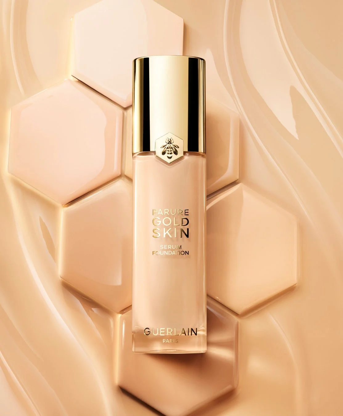 ゲランのPARURE GOLD SKIN SERUM FOUNDATIONのボトルが、ハニカム状のタイルと液体の質感を背景に配置された画像です。製品はゴールドのキャップとブランドを象徴するハチのエンブレムが特徴で、高級感のあるベージュトーンで統一されています。