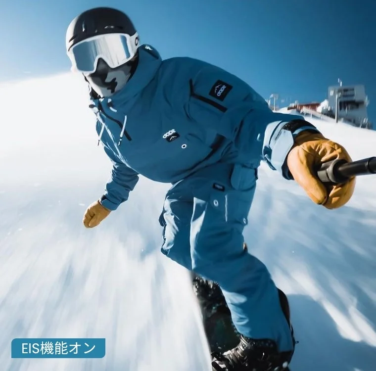 青いウェアを着た人物が、快晴の雪山をスノーボードで滑走している様子を捉えたダイナミックな写真です。画像下部には「EIS機能オン」の文字が見えます。