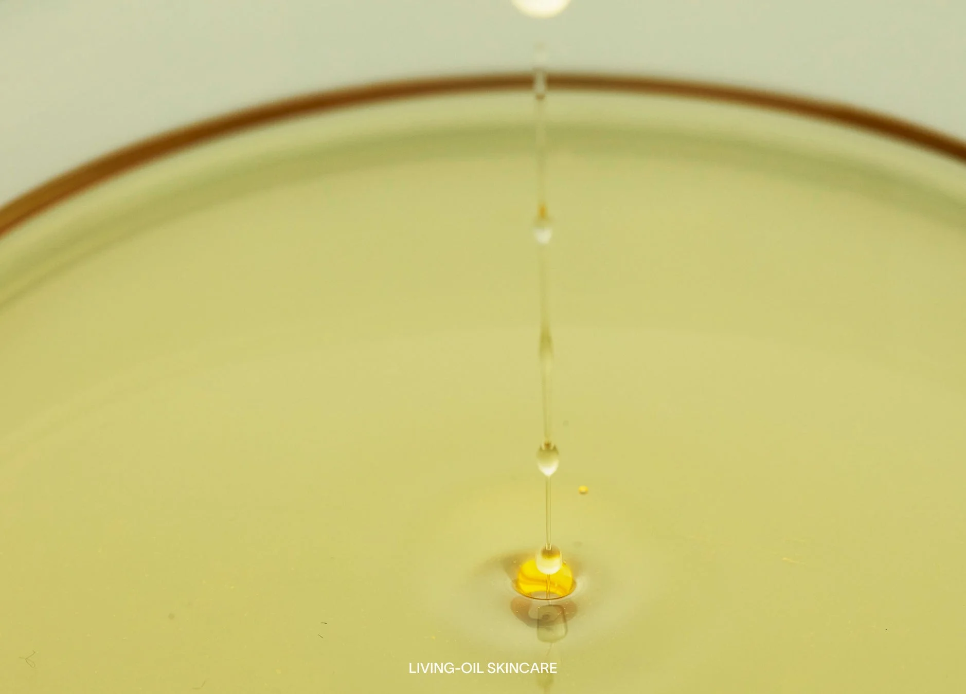 黄色い液体（オイル）が水面に滴り落ち、小さな波紋と飛び散るしずくが美しい瞬間を捉えています。自然な質感と滑らかさが感じられ、スキンケア製品を想起させます。