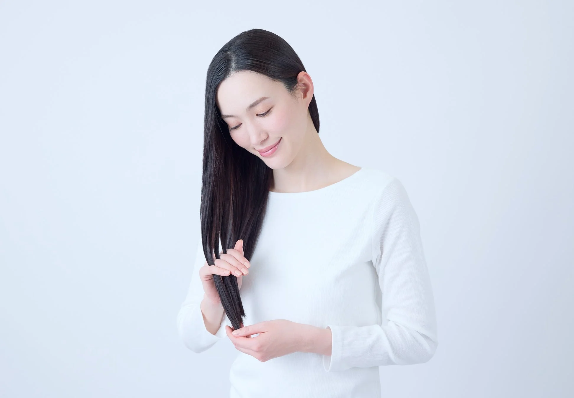 白い背景の前で、白い服を着た女性が微笑みながら自分の長く美しい黒髪を優しく触っています。
