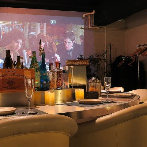 薄暗い照明のバーまたはレストランの店内で、壁に映画が映し出されています。カウンターには様々な種類の酒瓶、グラス、そして温かい光を放つキャンドルが並べられており、くつろいだ雰囲気を醸し出しています。