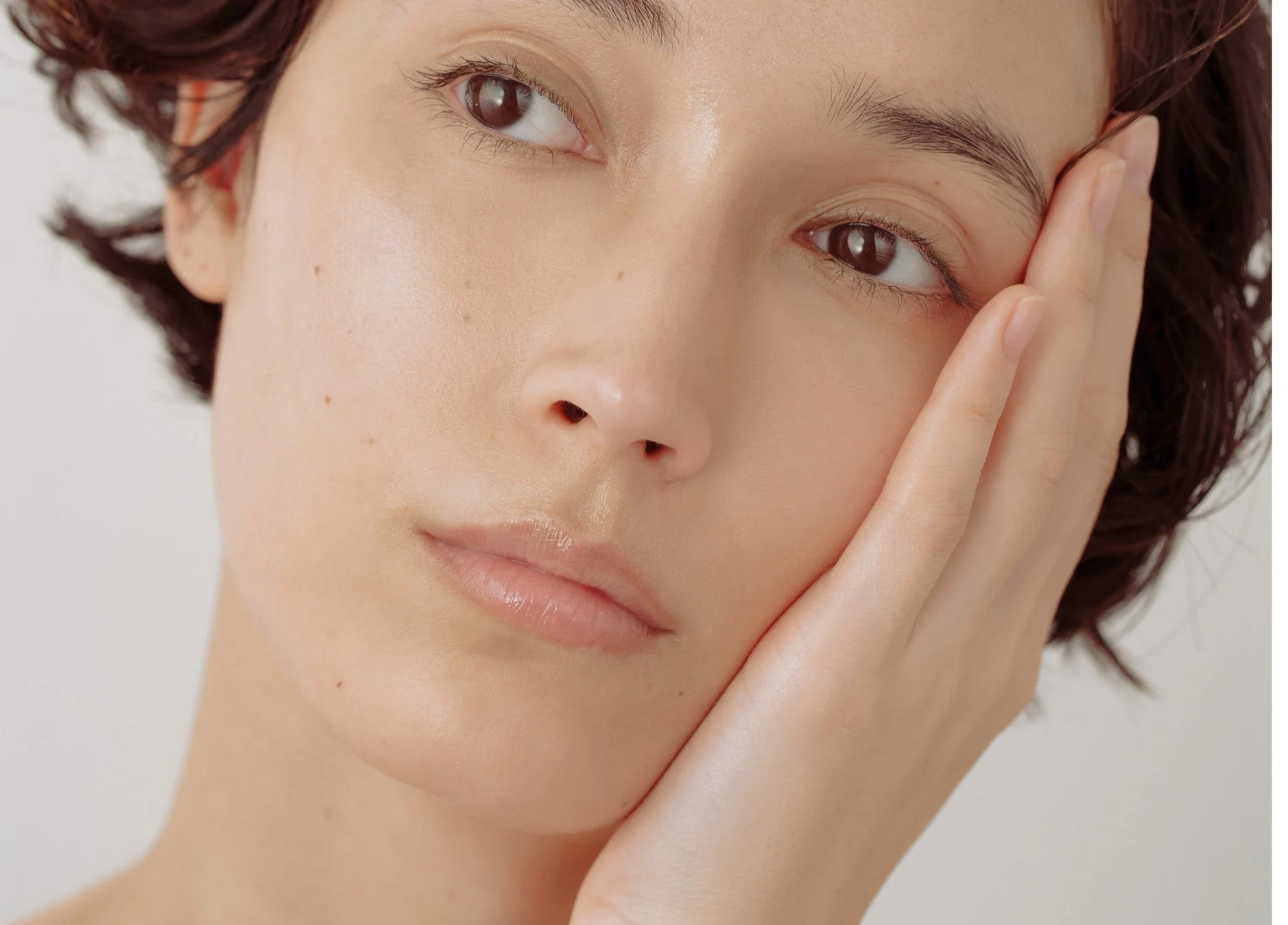透明感のある肌が美しい女性の顔のクローズアップ。片手を頬に添え、上を見つめるような表情が印象的です。自然な美しさが際立っています。