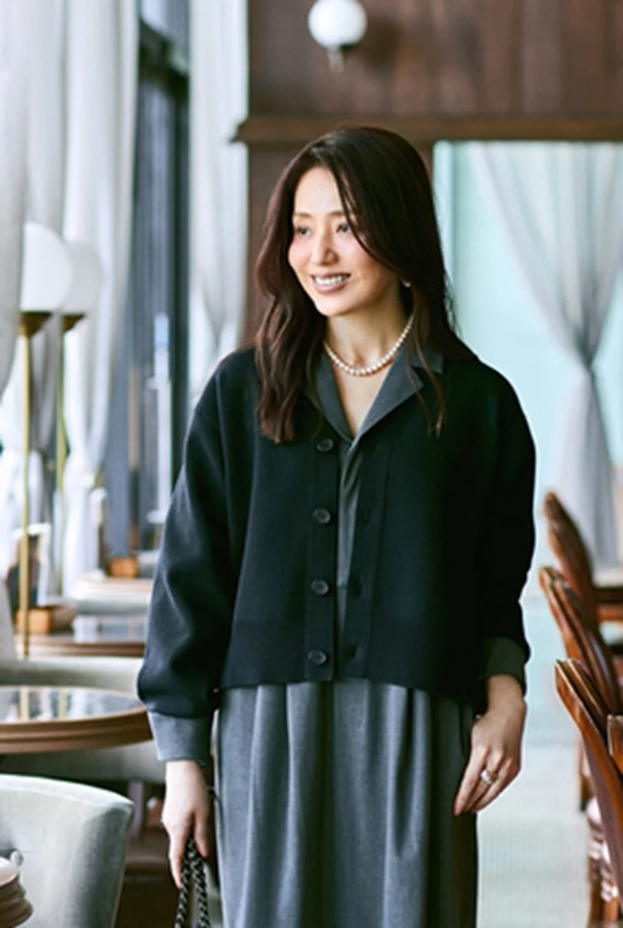 黒いカーディガンとグレーのワンピースを着用し、パールネックレスをつけた女性が笑顔で立っているポートレート。