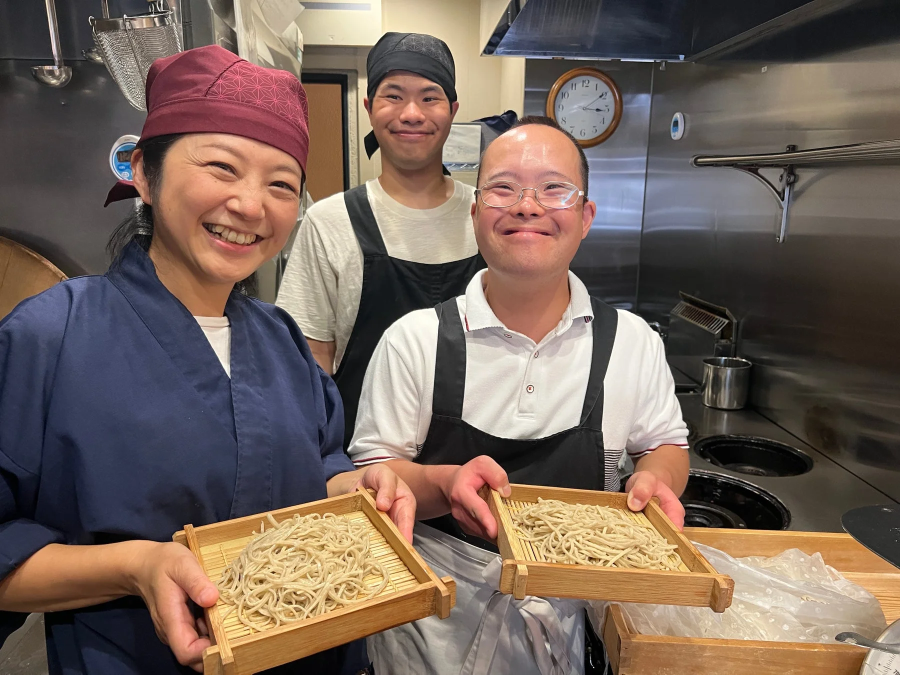 厨房で3人の人々が笑顔で手作りのそばが入った木製のざるを持っている