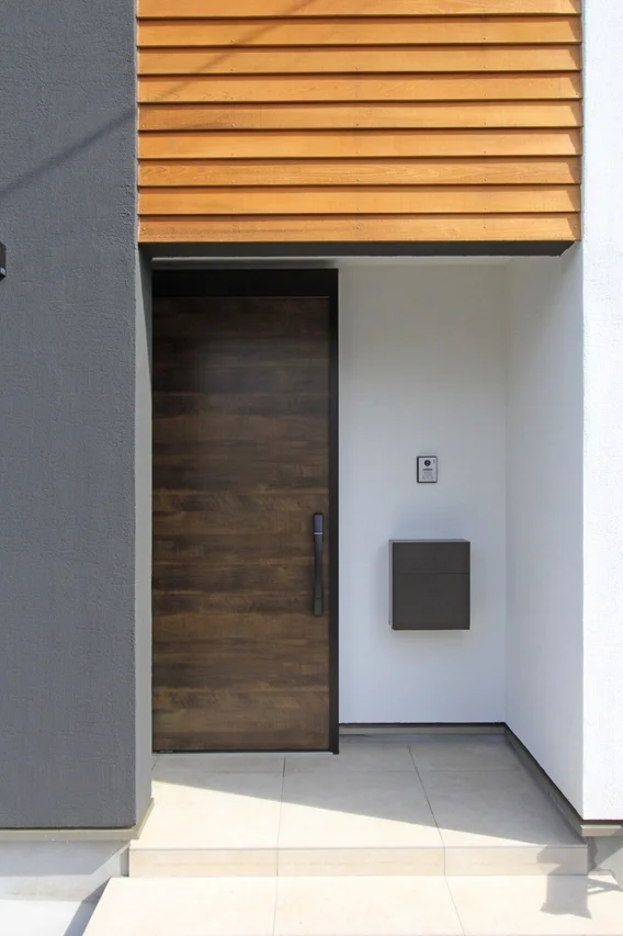 木目調のドアと木製ルーバーの天井が特徴的なモダンな住宅の玄関です。グレーと白の壁に囲まれ、シンプルなポストとインターホンが設置されています。