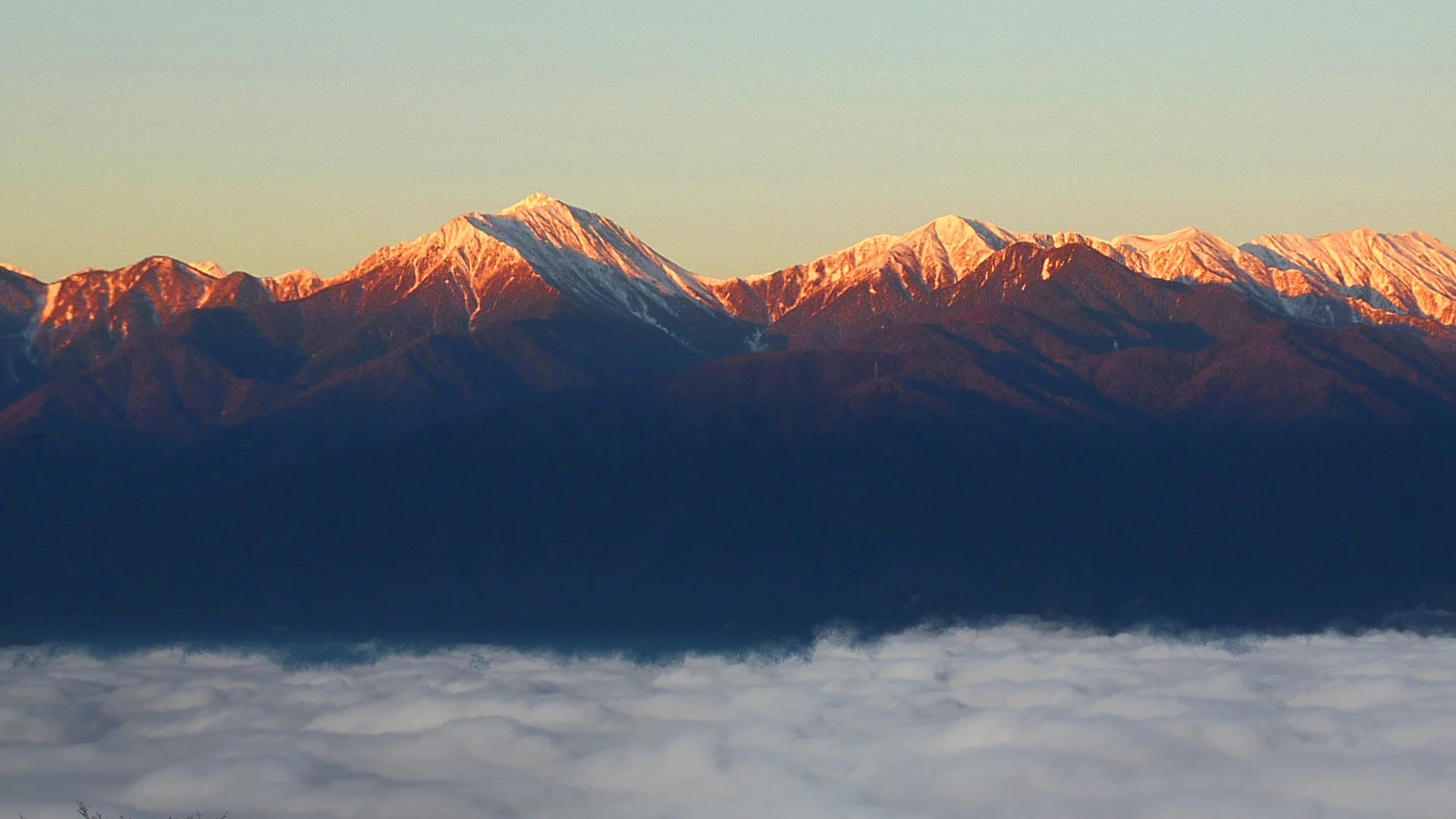 雪を冠した雄大な山々が朝日に照らされ、山頂が黄金色に輝いています。手前には広大な雲海が広がり、幻想的な自然の風景が広がっています。