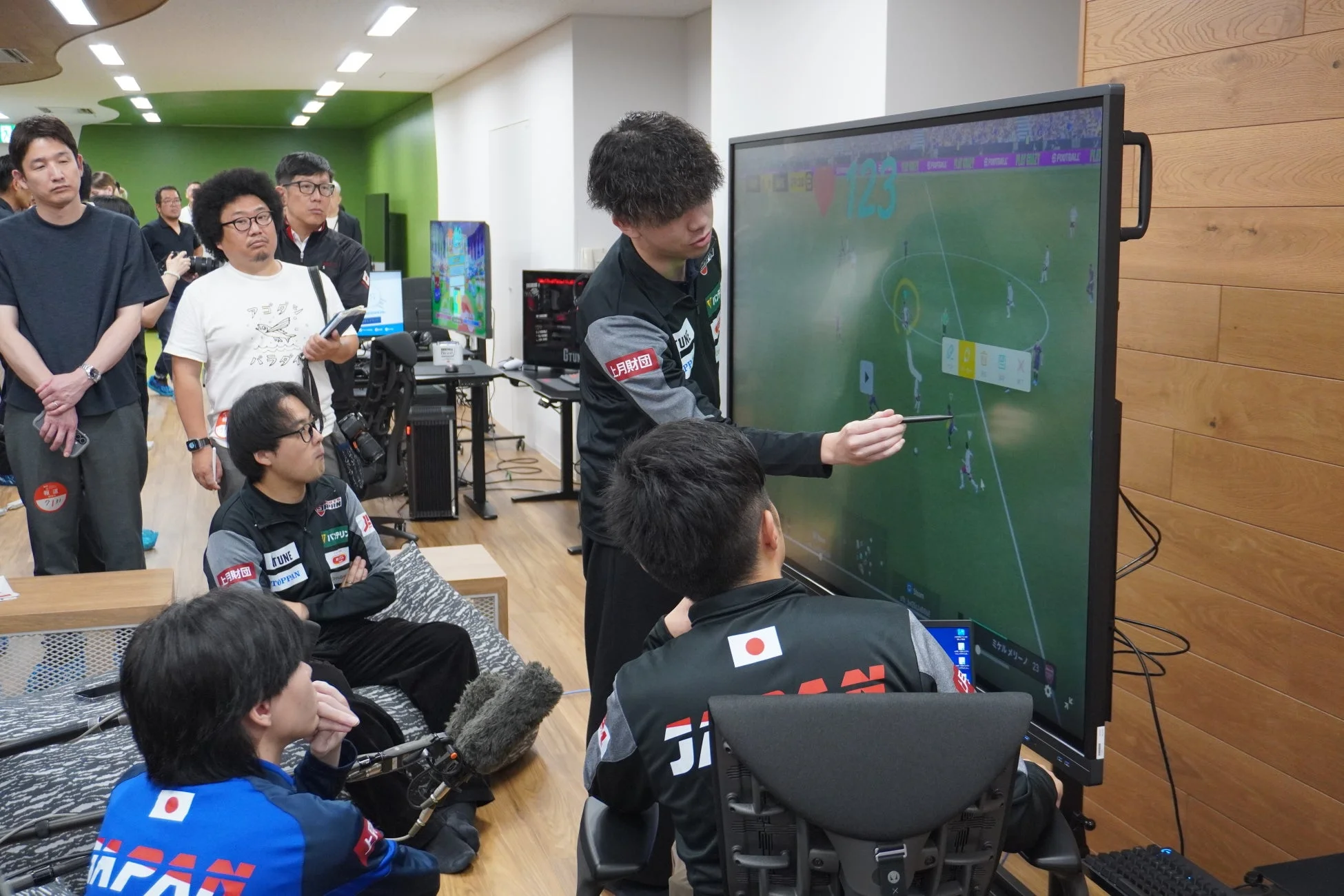 eスポーツのサッカーゲームをプレイする選手たちが、大画面モニターを見ながら戦略を話し合っている様子